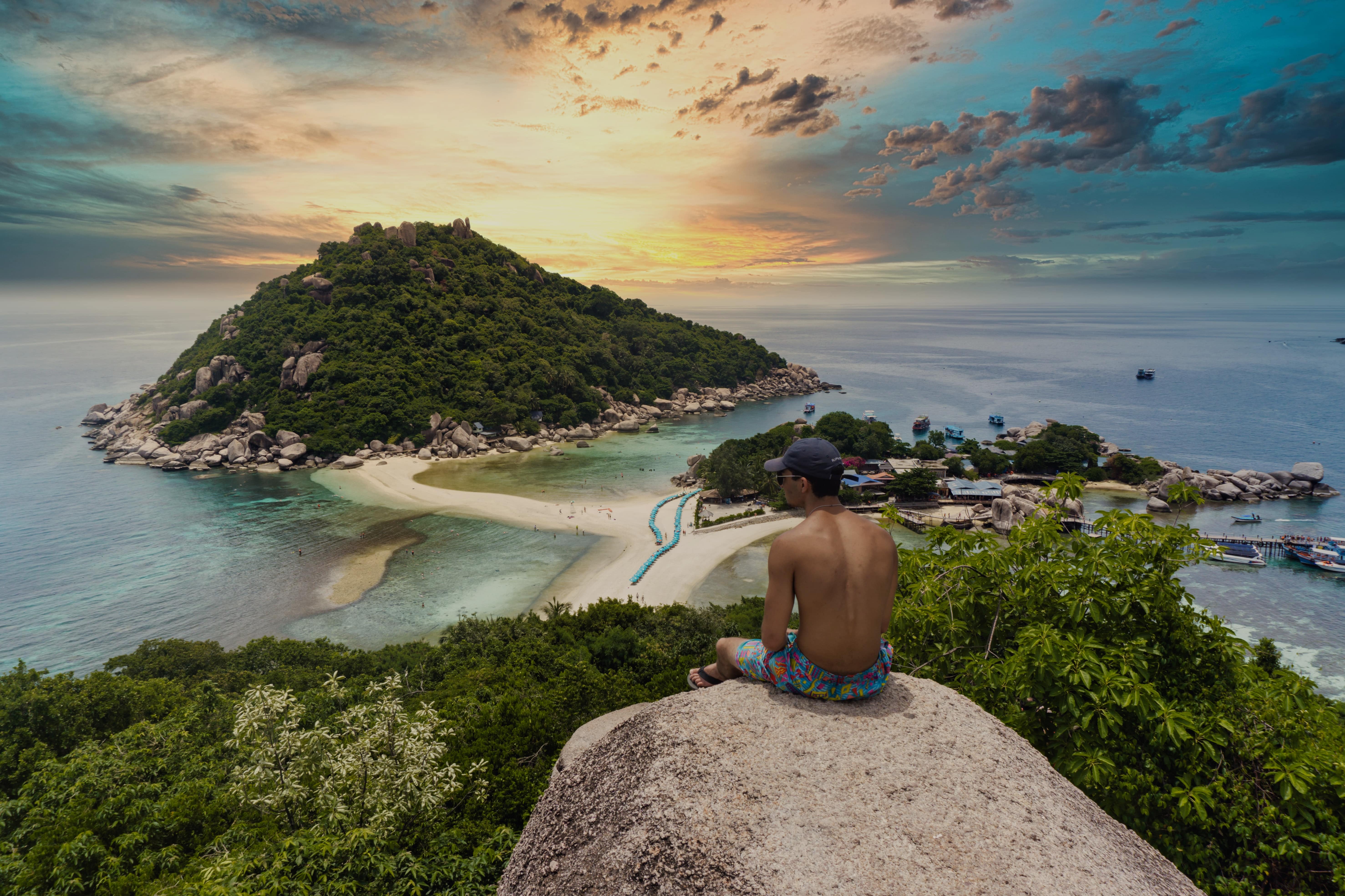 Person sitting on a cliff overlooking a Thai bay, symbolizing ThaiTourConnect's mission to connect Thai tours with the online community.